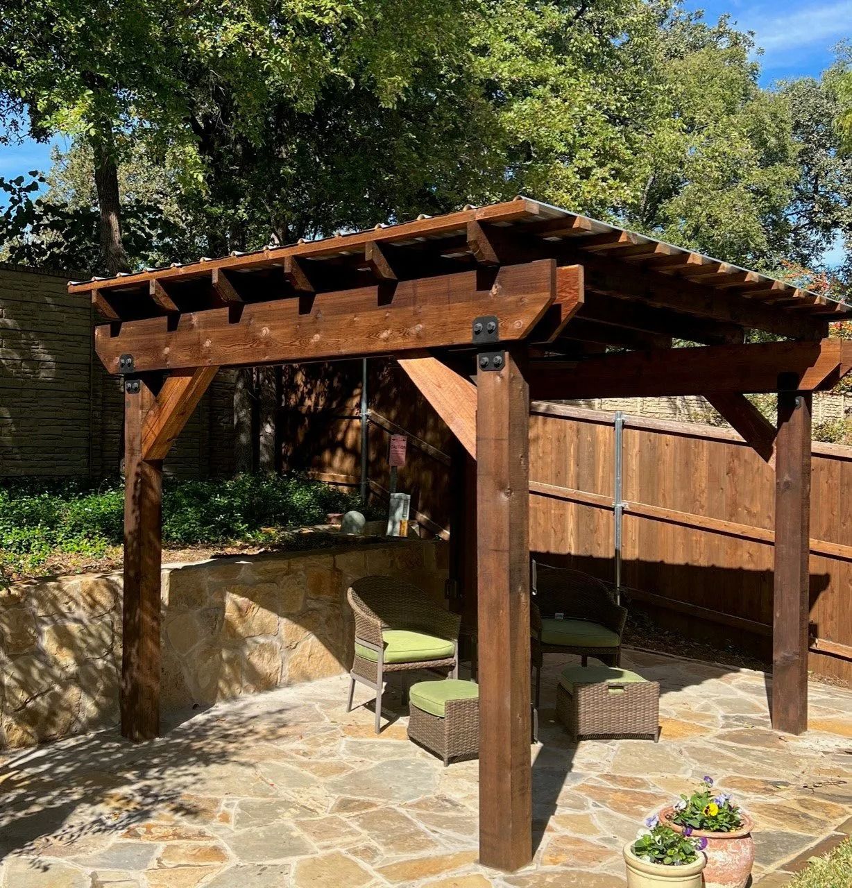 Wooden pergola over a stone patio with chairs, in a sunny backyard garden