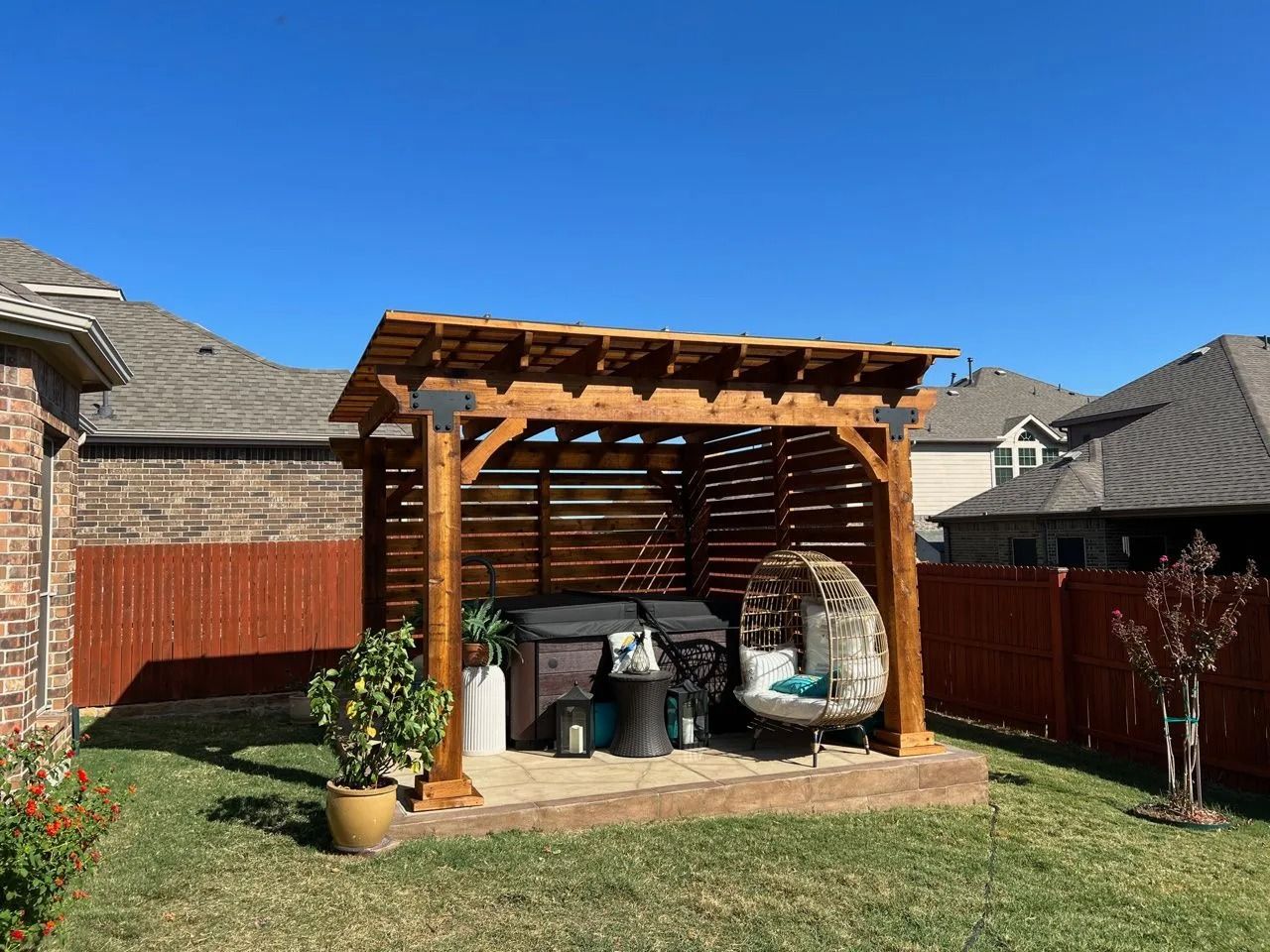 Wooden backyard pergola sheltering patio furniture and grill beside a fenced lawn under a clear blue sky