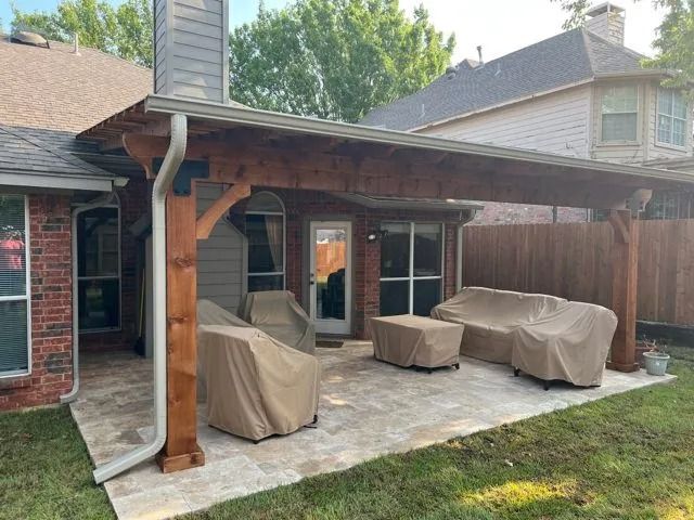 Covered patio with two tan-covered outdoor chairs and a matching table beside a brick house and wooden fence.
