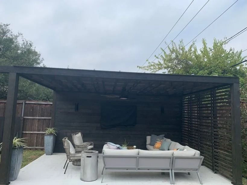 Covered patio with outdoor sofa and chairs beneath a dark pergola, overlooking a backyard.