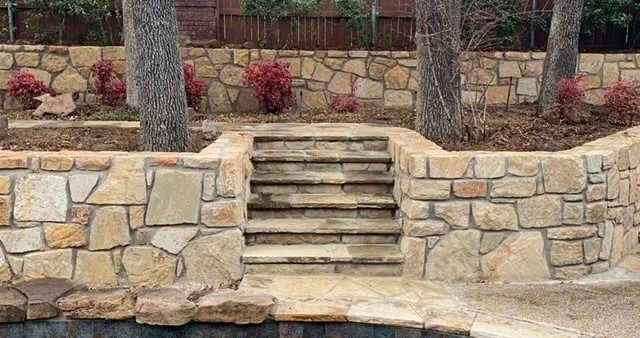 Stone steps built into a low retaining wall in a landscaped yard with shrubs and trees.