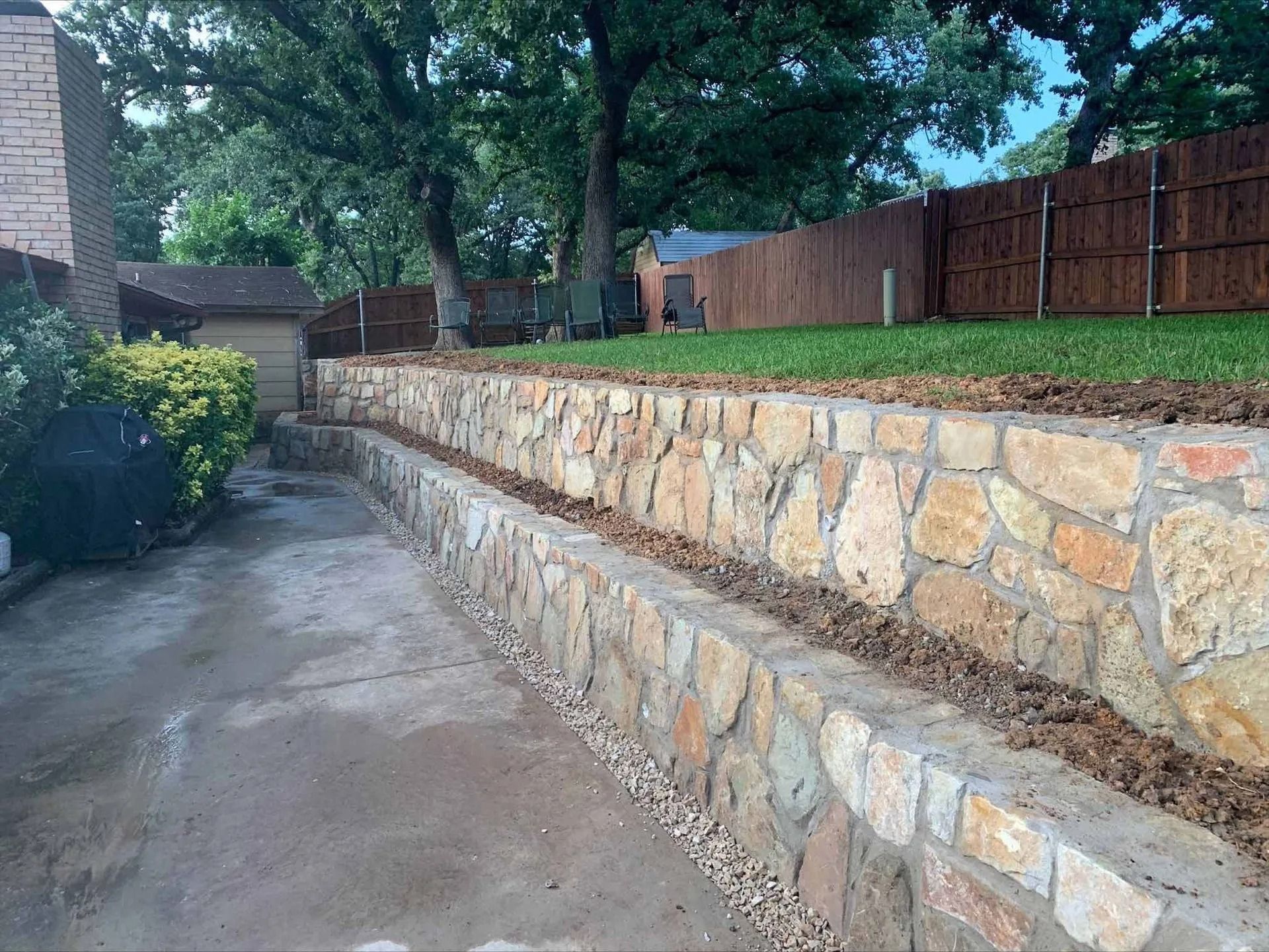 Stone retaining wall along a paved driveway with trees and a wooden fence in the background