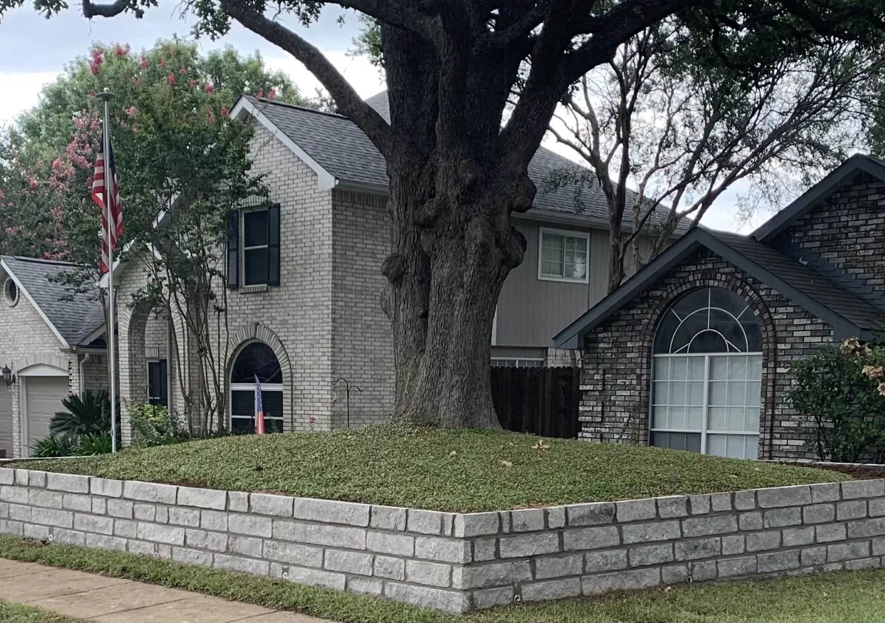 Stone house with a large tree and low brick retaining wall, seen from the street.