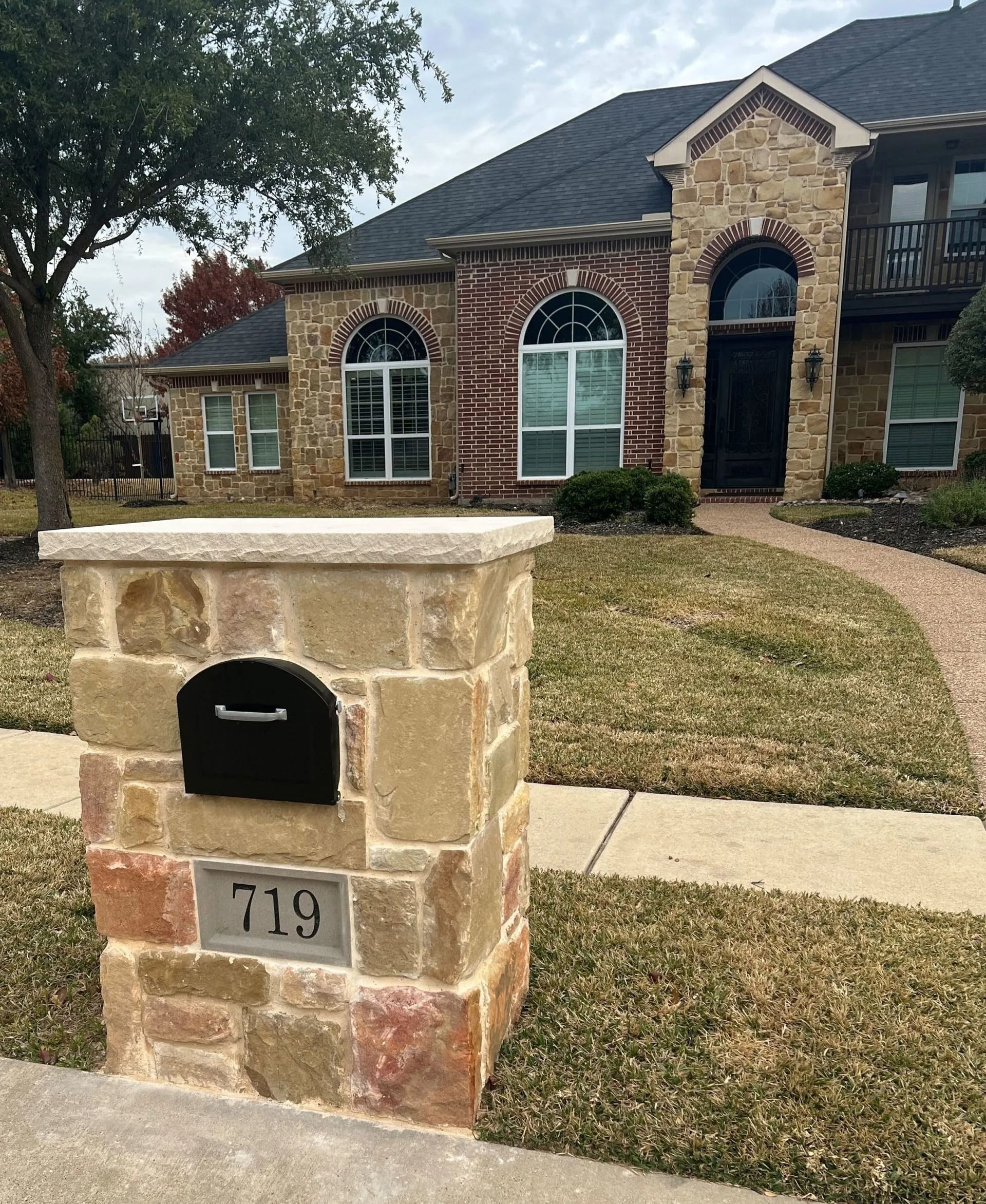 Stone mailbox in front of a brick house with arched windows and a walkway; house number 719.