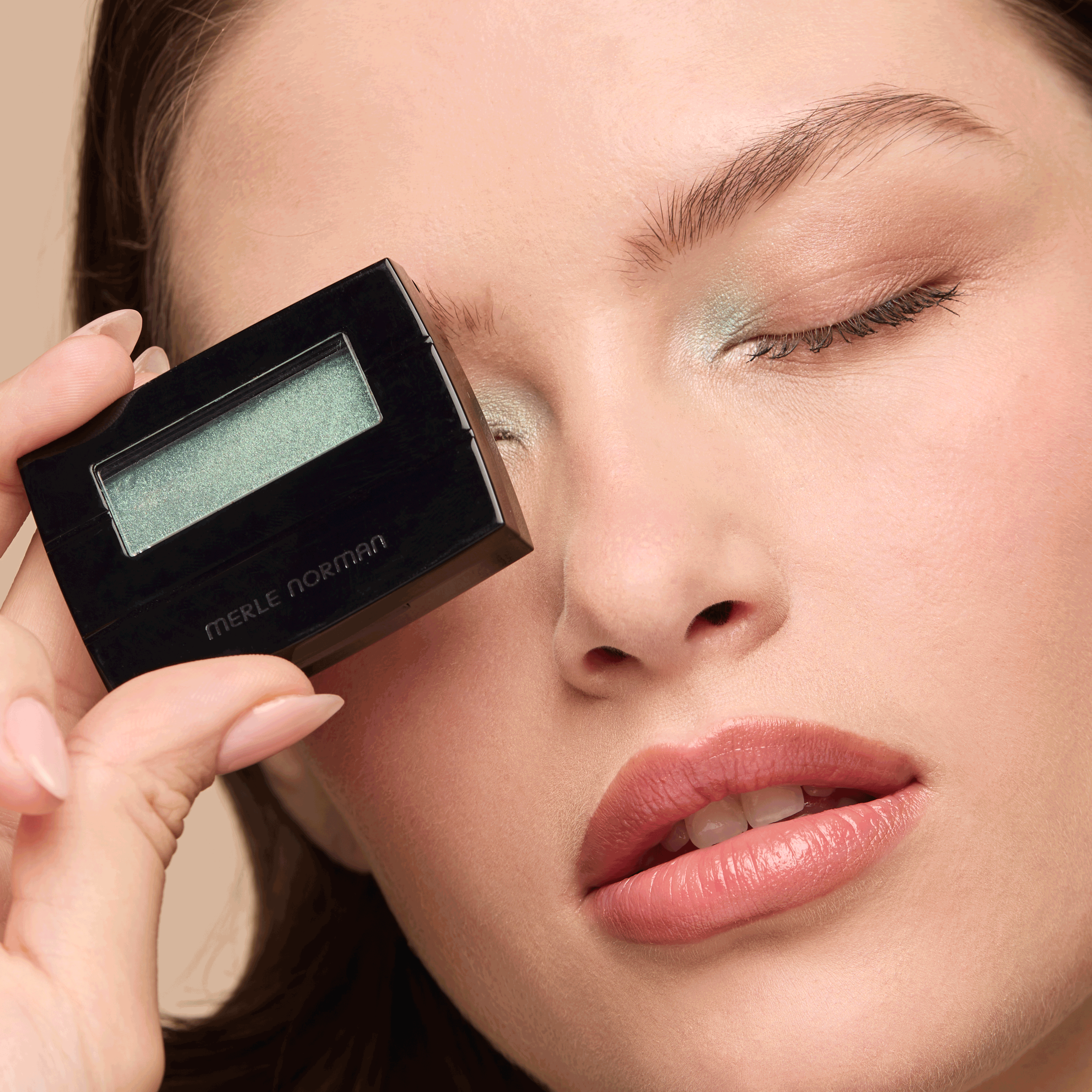 A person holds a small, black eyeshadow compact with shimmery light green shadow next to their eye, which matches the shade