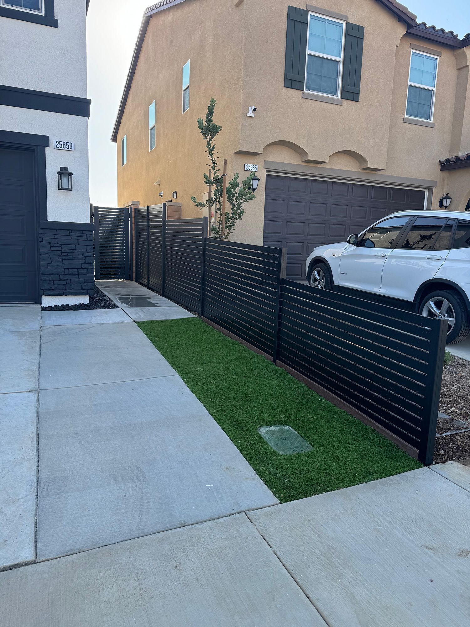 Black slatted fence lines a walkway next to a tan house with a white car in the driveway.