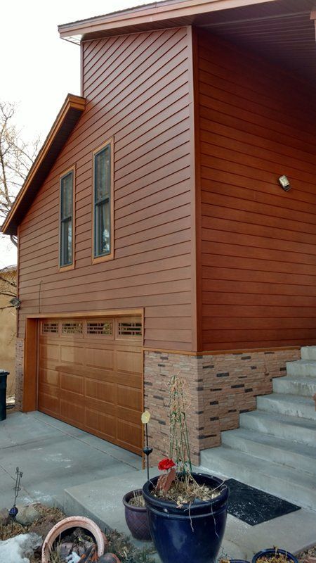 Brown siding on a two-story house with a garage and brick base. A few potted plants sit on the concrete.