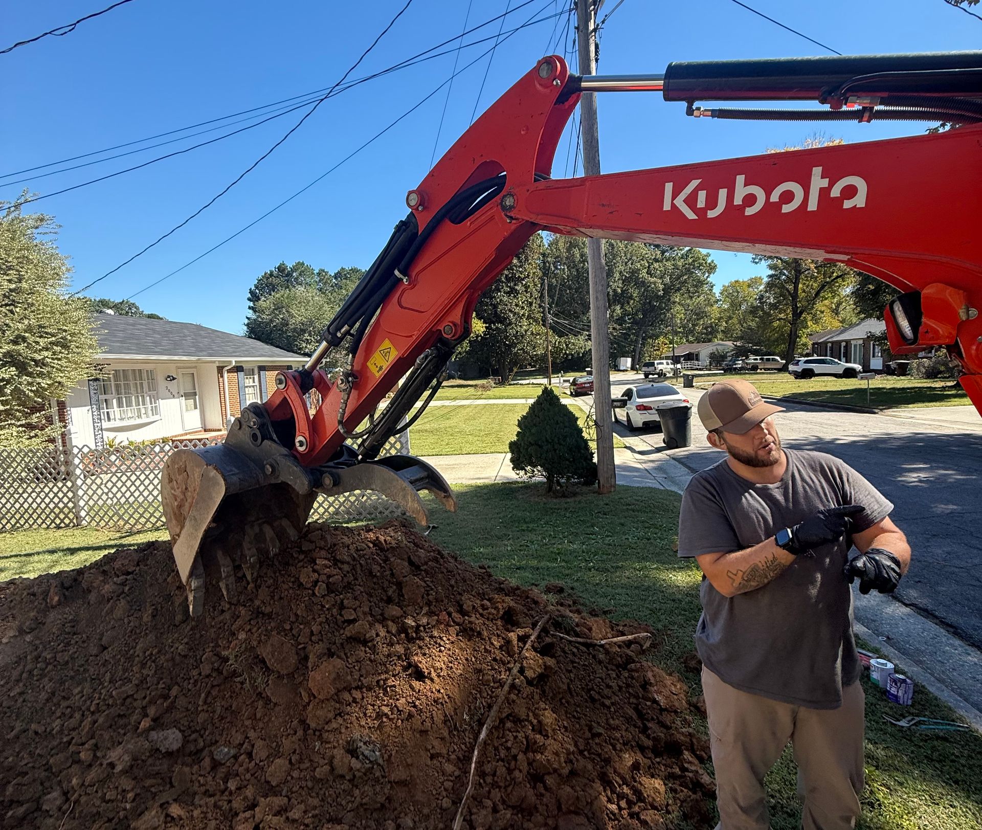 Man watches a red Kubota excavator digging dirt in a residential yard on a sunny day.