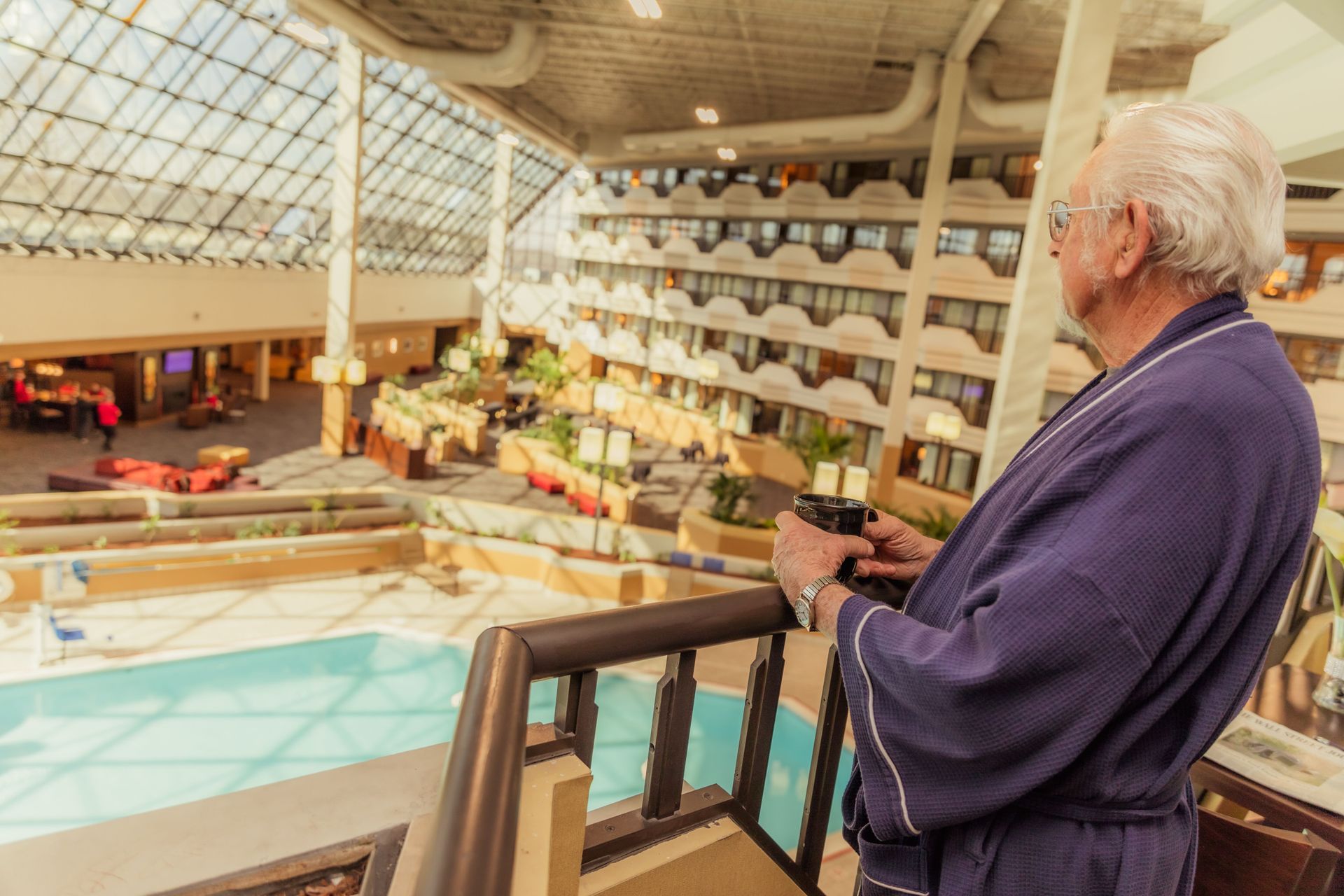 An older man in a bathrobe is standing on a balcony overlooking a swimming pool.