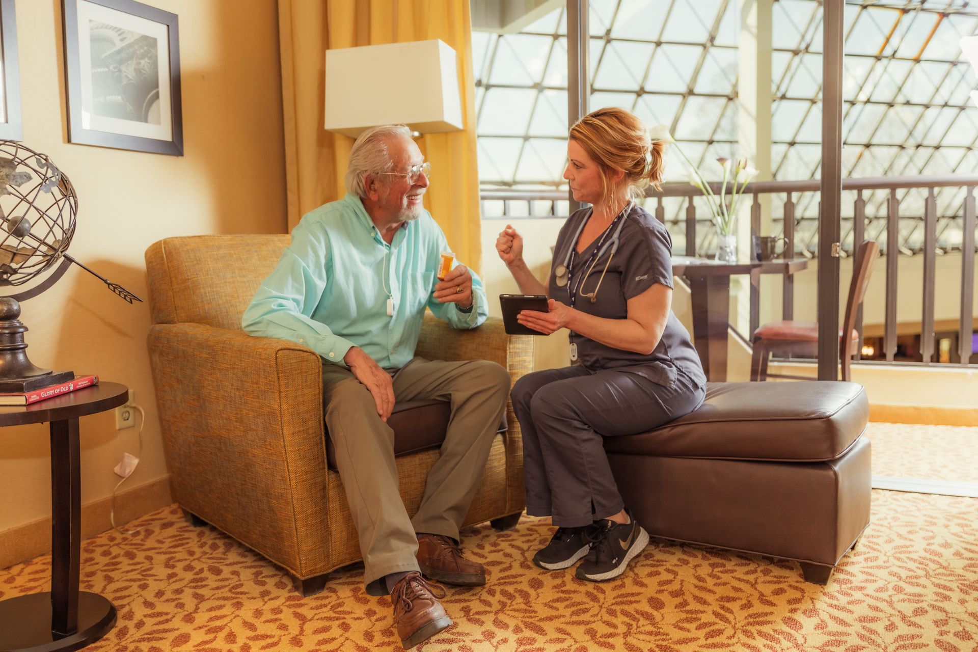A nurse is sitting on an ottoman talking to an elderly man in a chair.