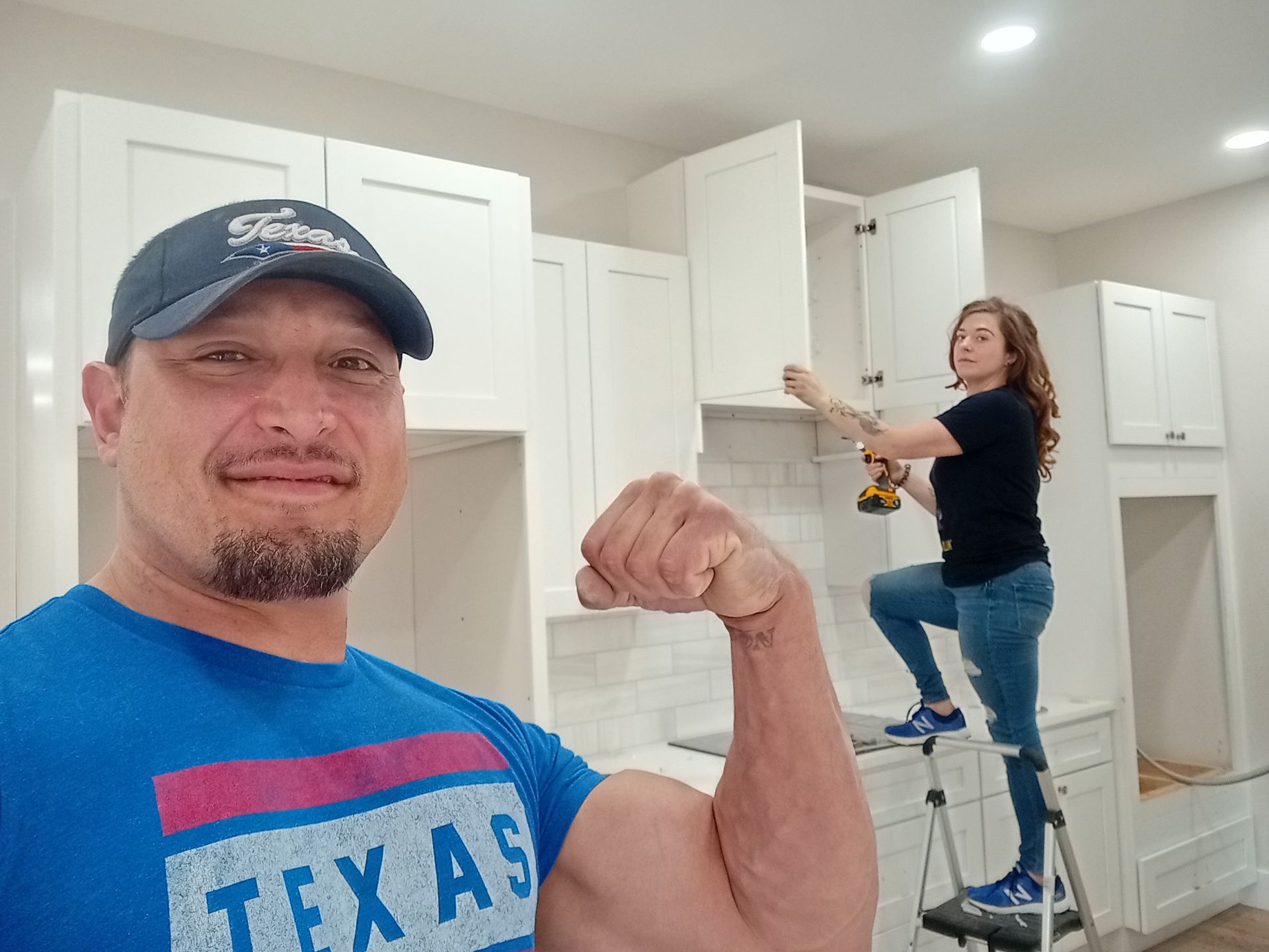 A man is flexing his muscles in a kitchen while a woman stands on a ladder.