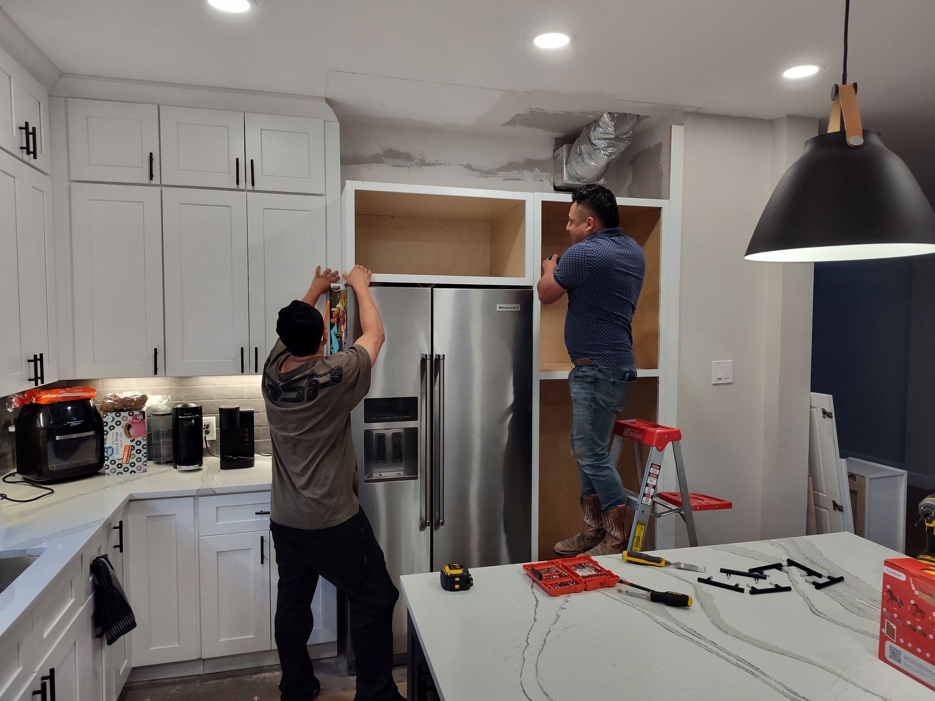 Two men are working on a refrigerator in a kitchen.