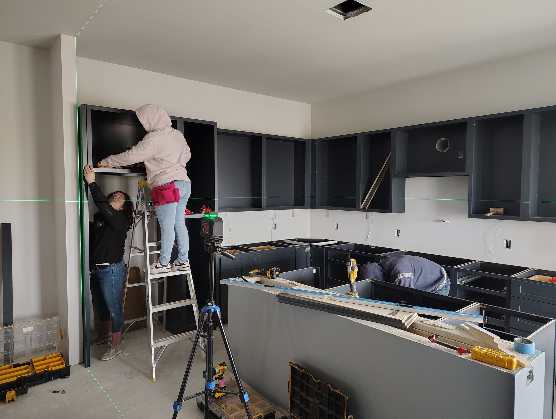 A woman is standing on a ladder measuring a cabinet in a kitchen.