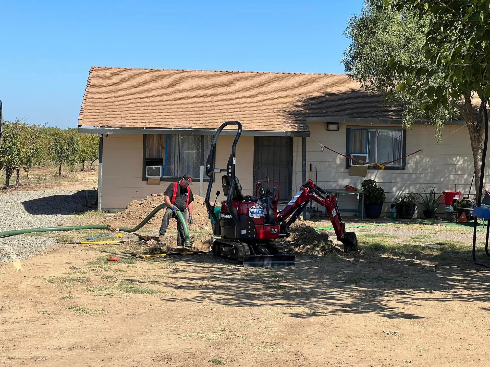 Person using an excavator to dig near a small, beige house with a brown roof. Vineyard in the background.