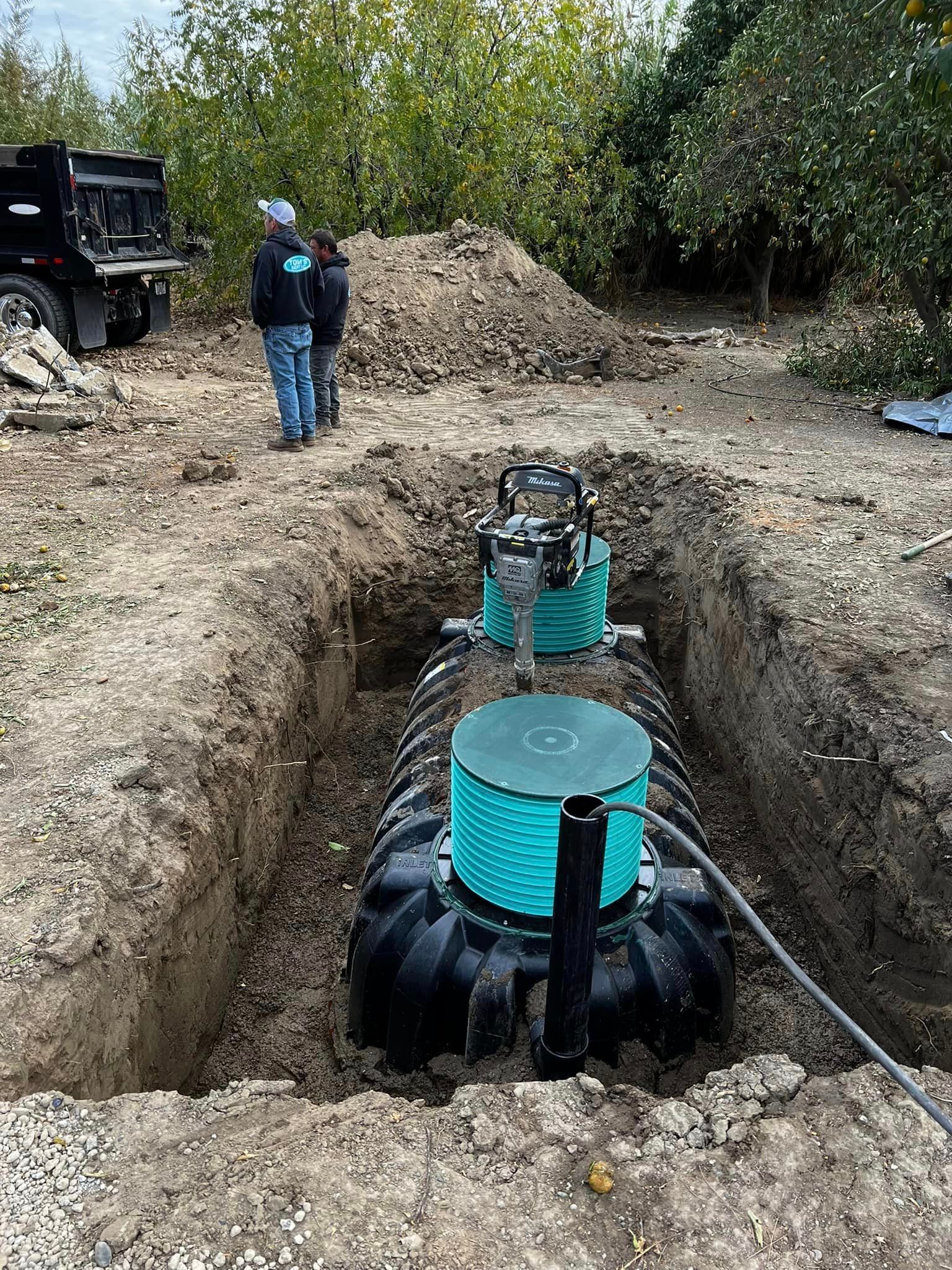 Septic tank installation: Black tank in a trench, two workers, equipment, and a dump truck on site.