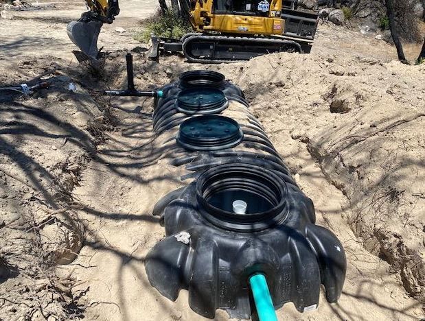 Black septic tank installed in a sandy trench; small excavator in the background.