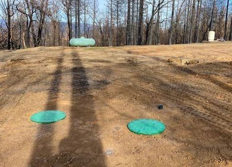 Two green septic tank lids in a dirt clearing with a propane tank and trees in the background.