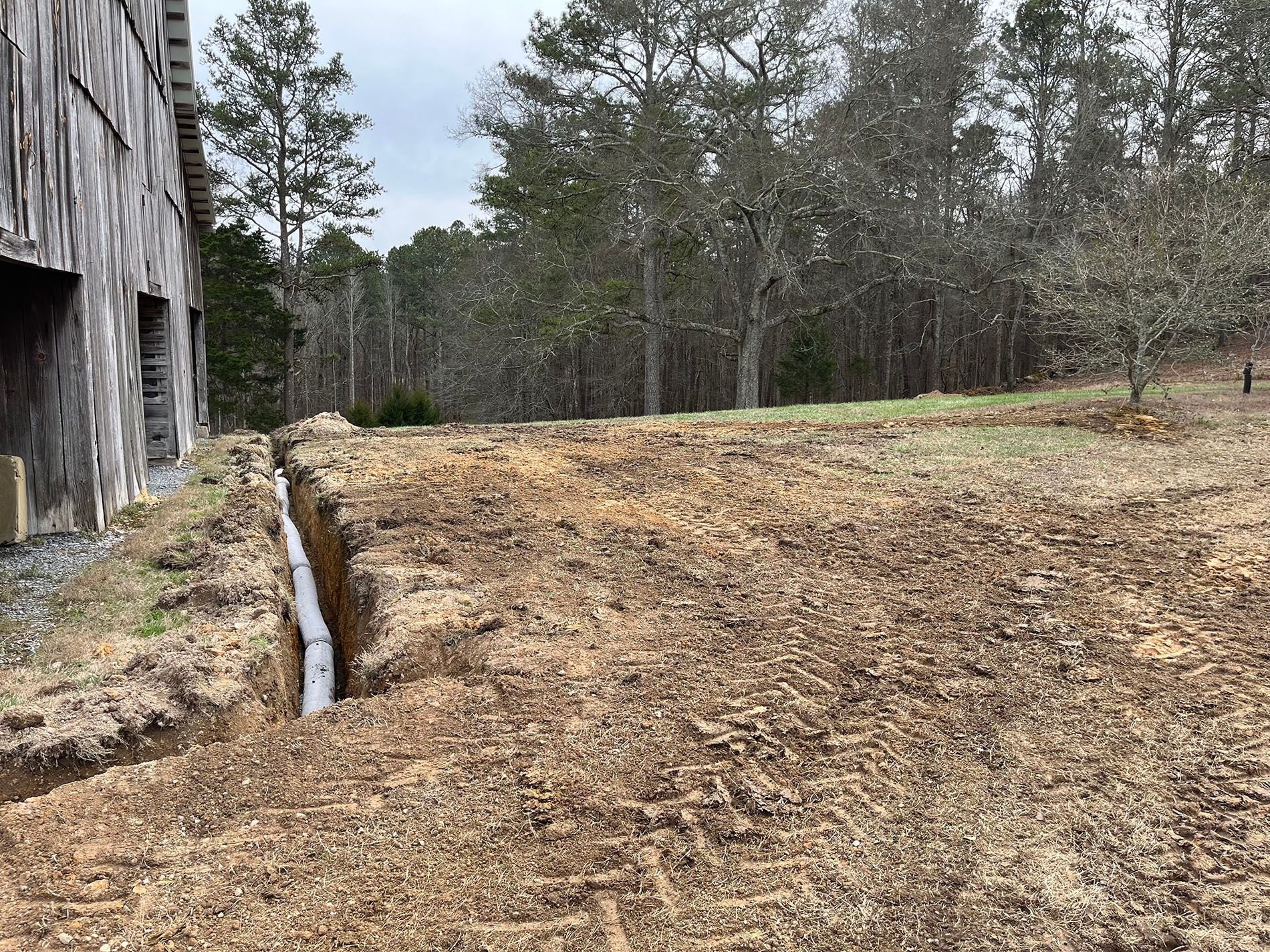A drainage pipe is being installed in a dirt field in front of a barn.