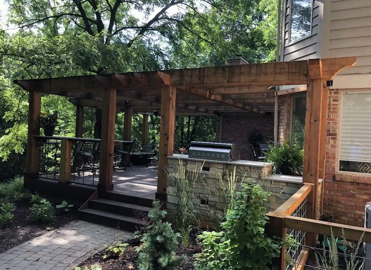 Wooden pergola over a patio with a grill and outdoor seating, surrounded by trees.