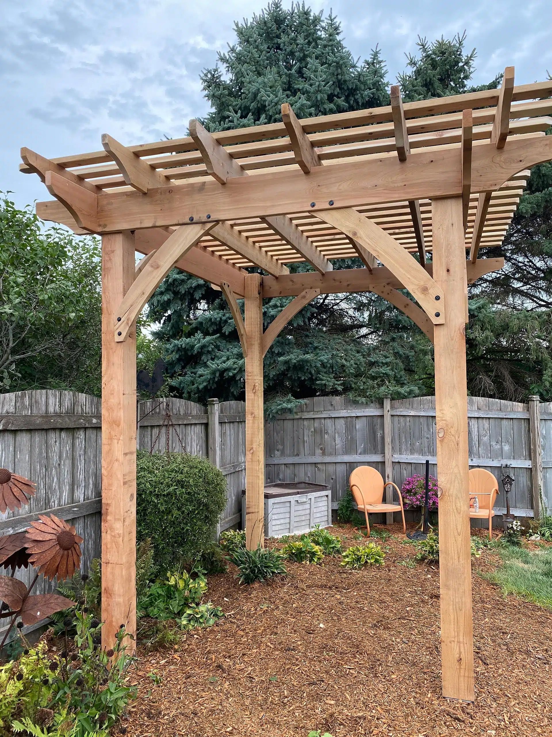 Wooden pergola in a backyard garden with chairs, mulch, and wooden fence.