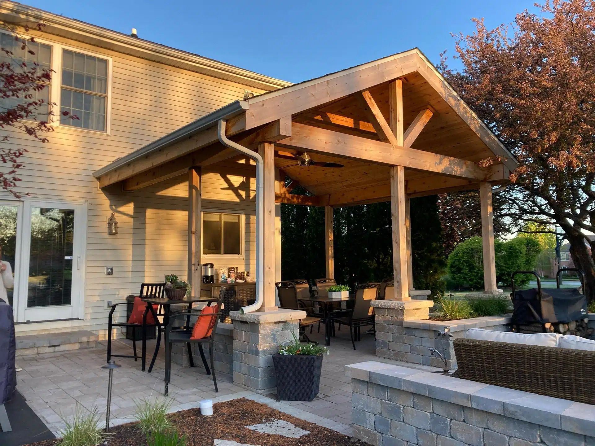 Outdoor patio with wooden roof extension, stone columns, dining table, and landscaping.