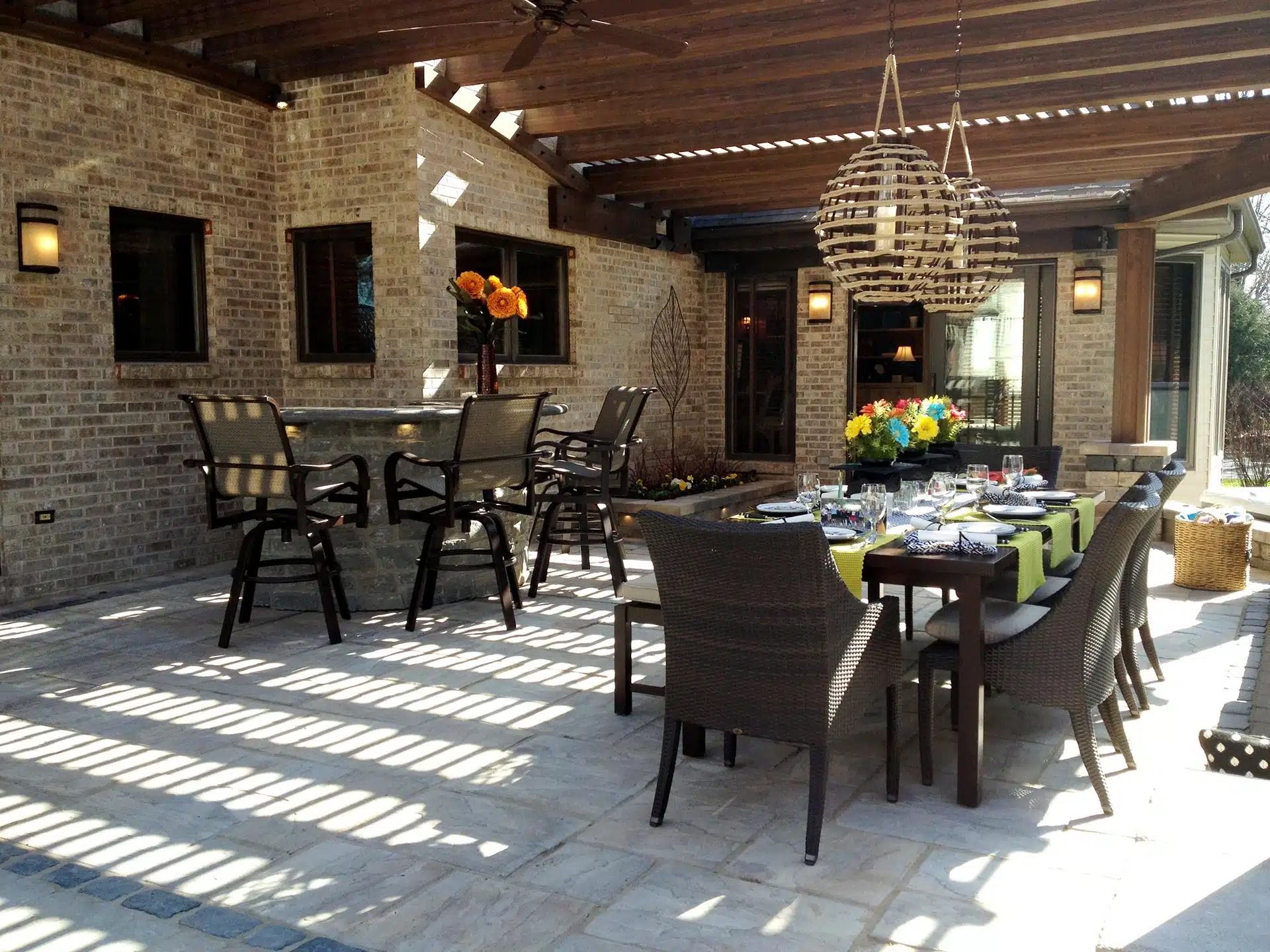 Outdoor patio with dining table and bar seating under a wooden pergola, brick walls, sunlight creating shadows.