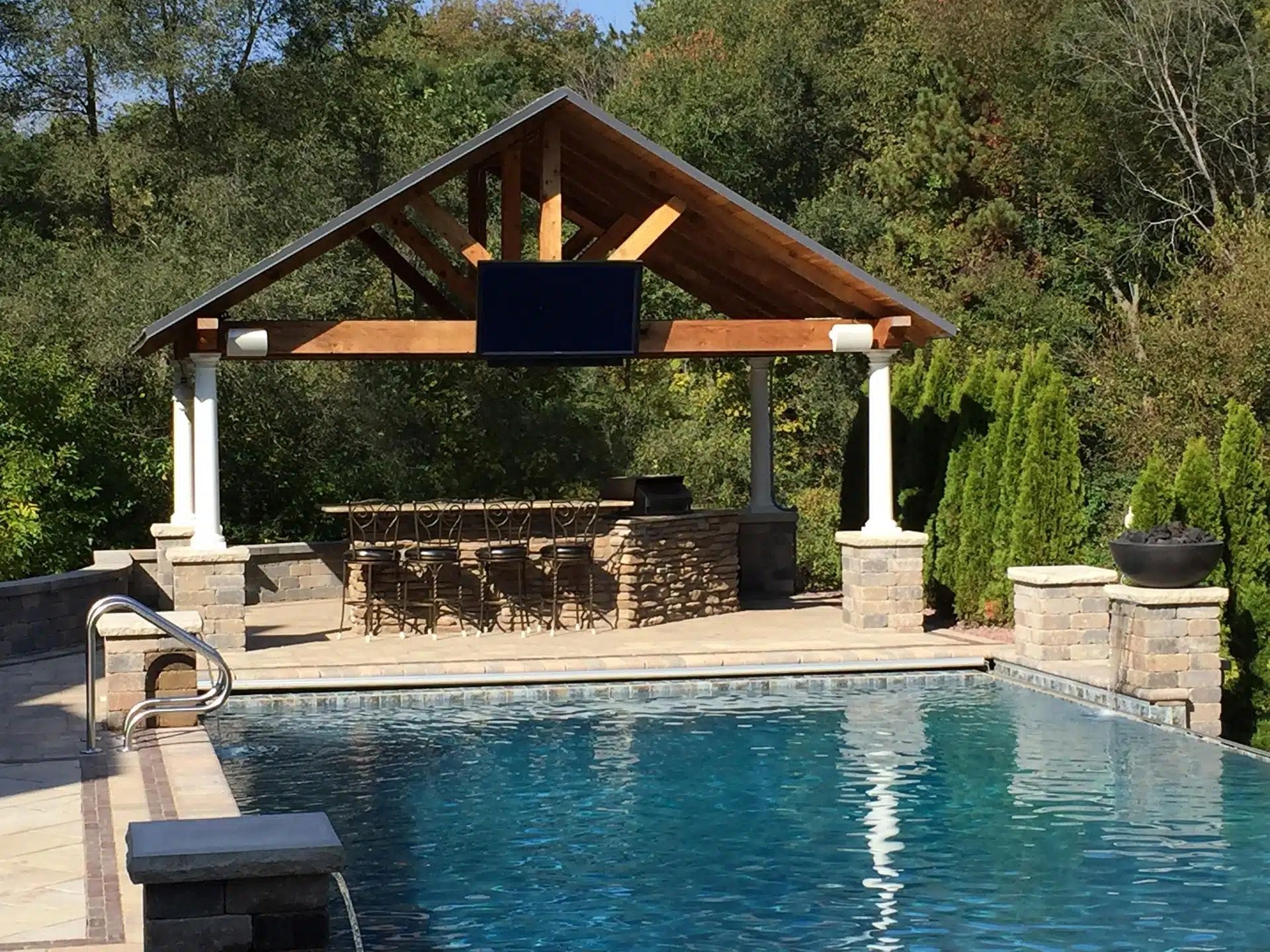 Poolside pavilion with TV, bar, and seating, overlooking a swimming pool.