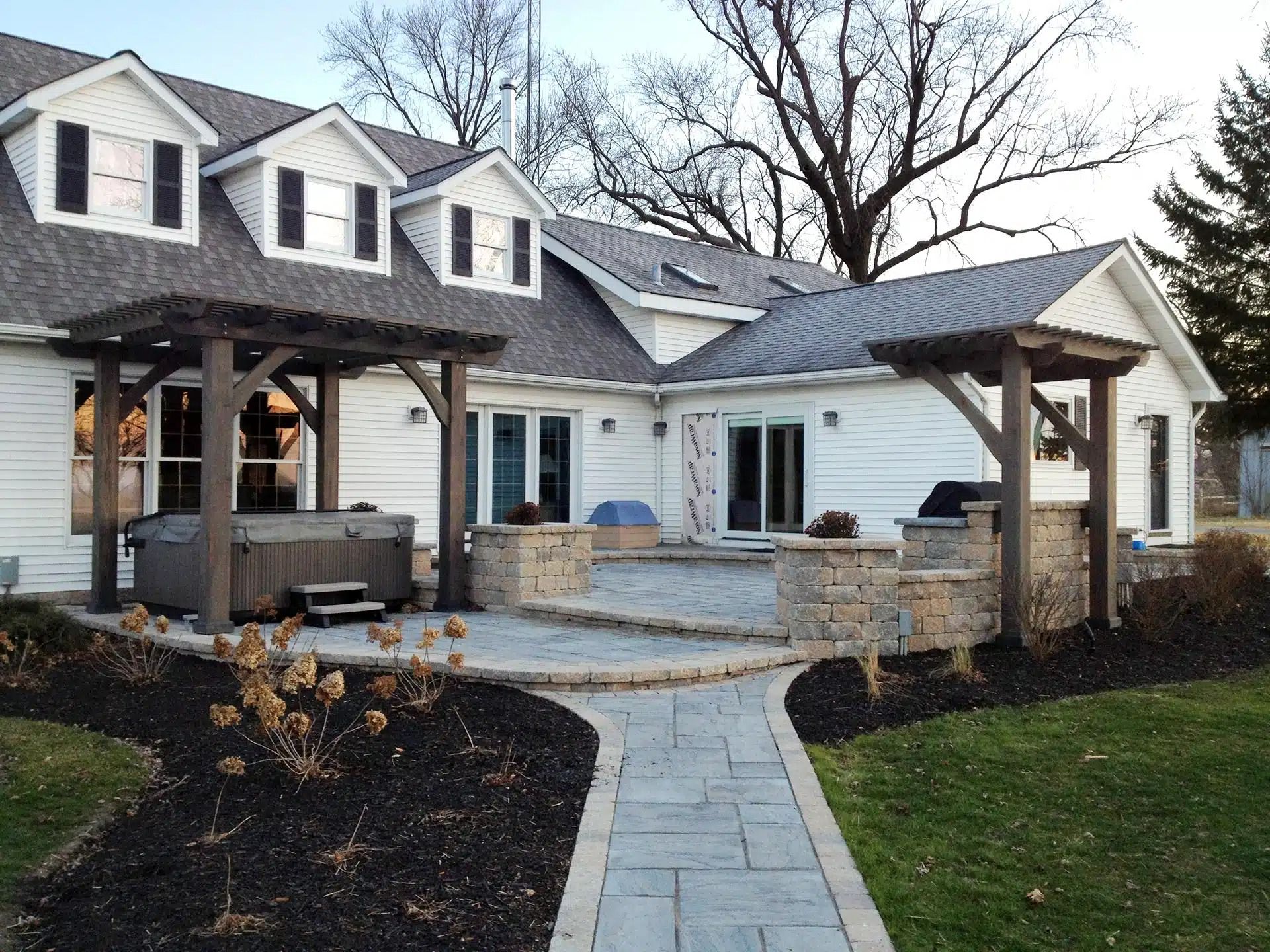 Backyard patio with a hot tub, stone walls, and wooden pergolas; a white house in the background.