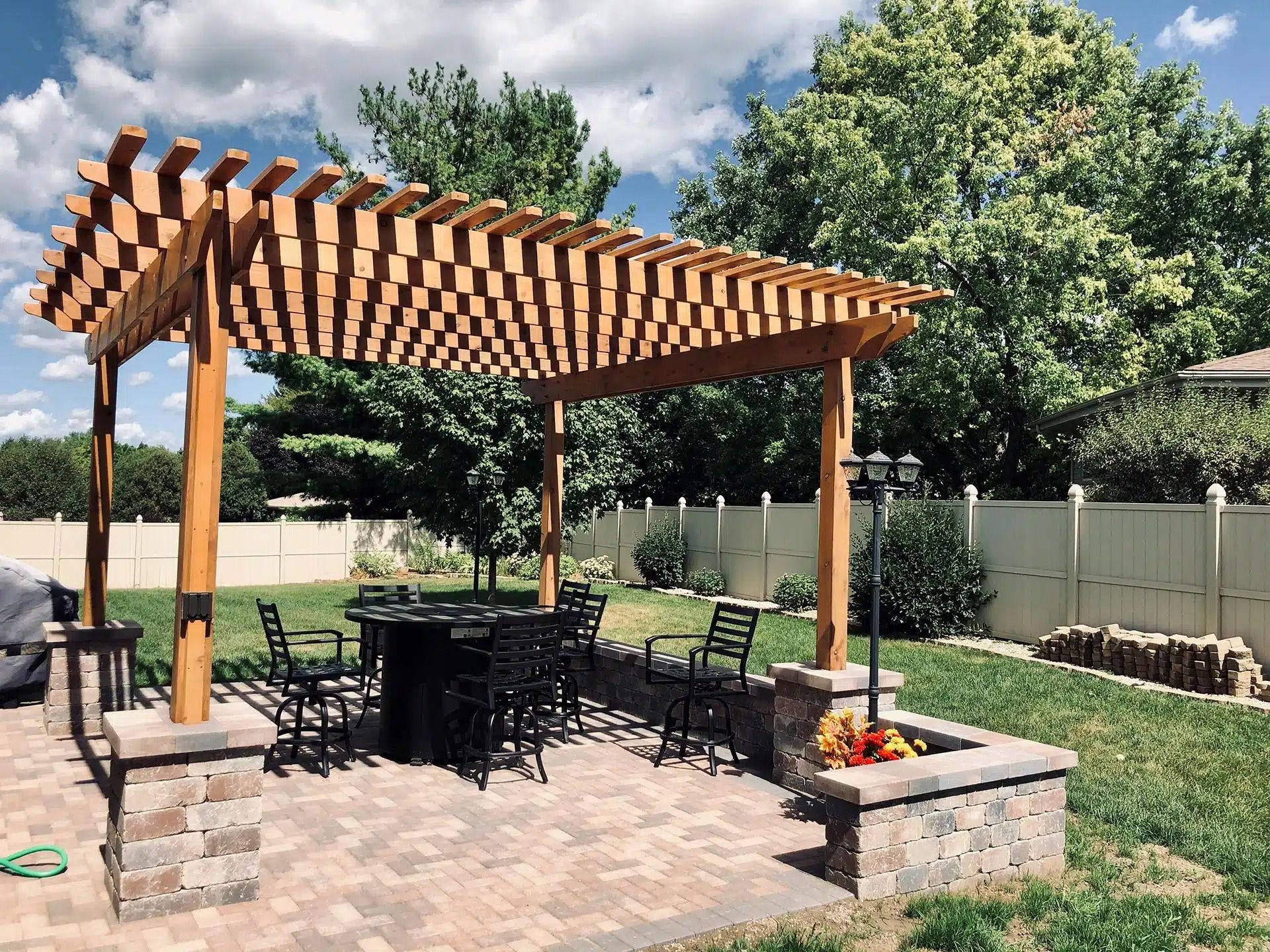 Wooden pergola over patio with table and chairs. Fire pit and white fence in background.
