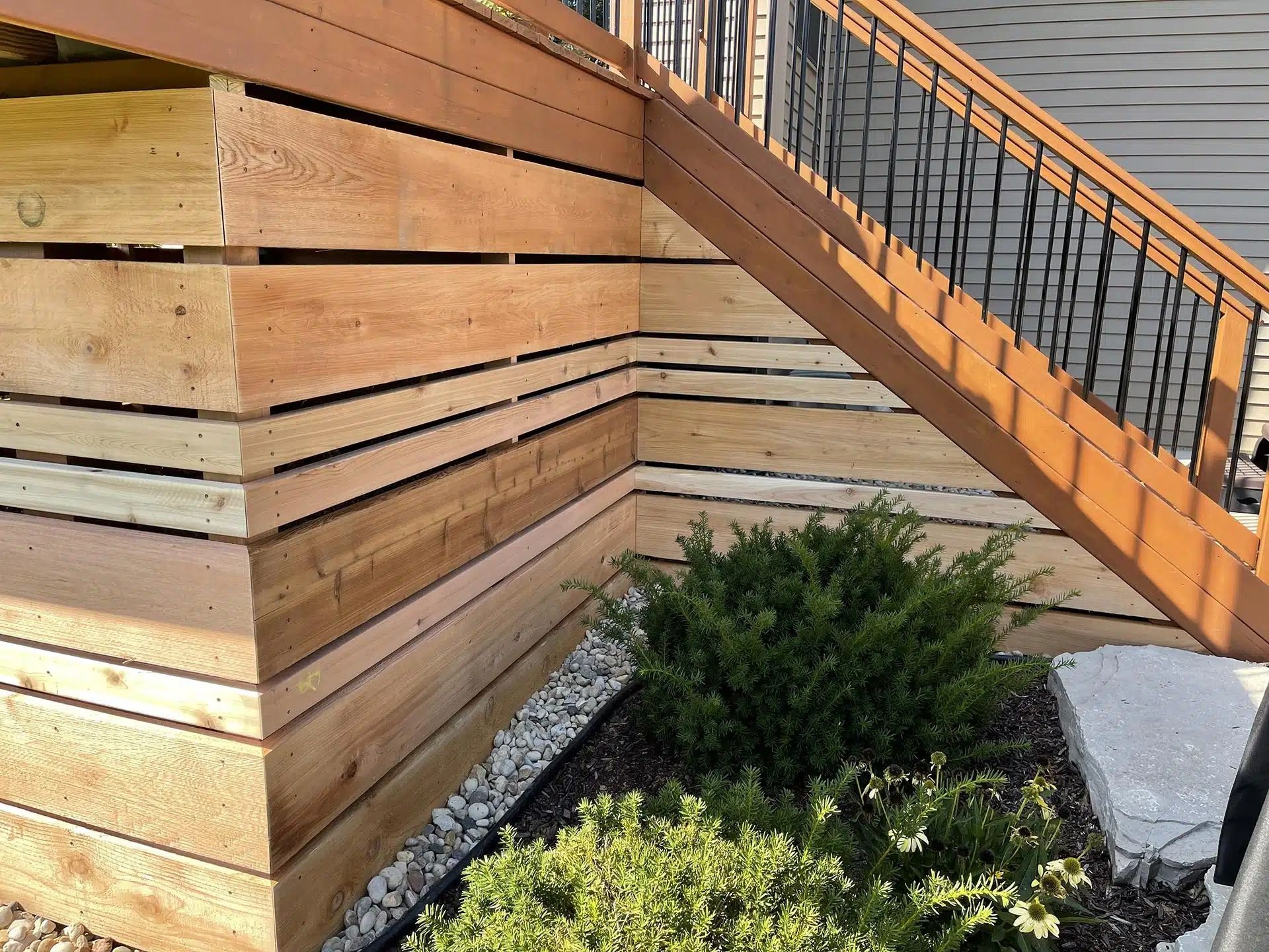 Wooden deck and stairs with black railing over a landscaped area with a bush and rock border.