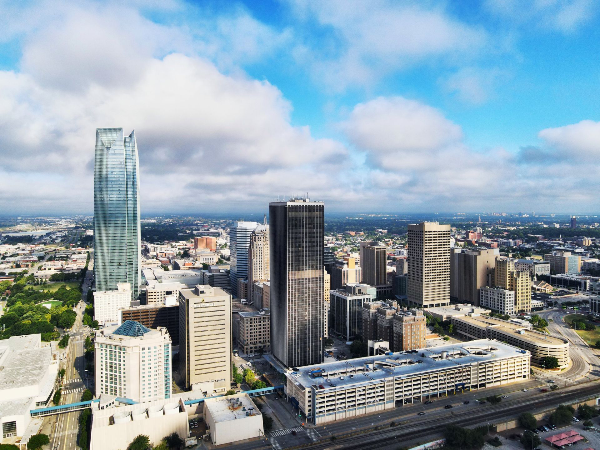 City skyline with modern high-rises, a tall glass tower, and a cloudy blue sky.