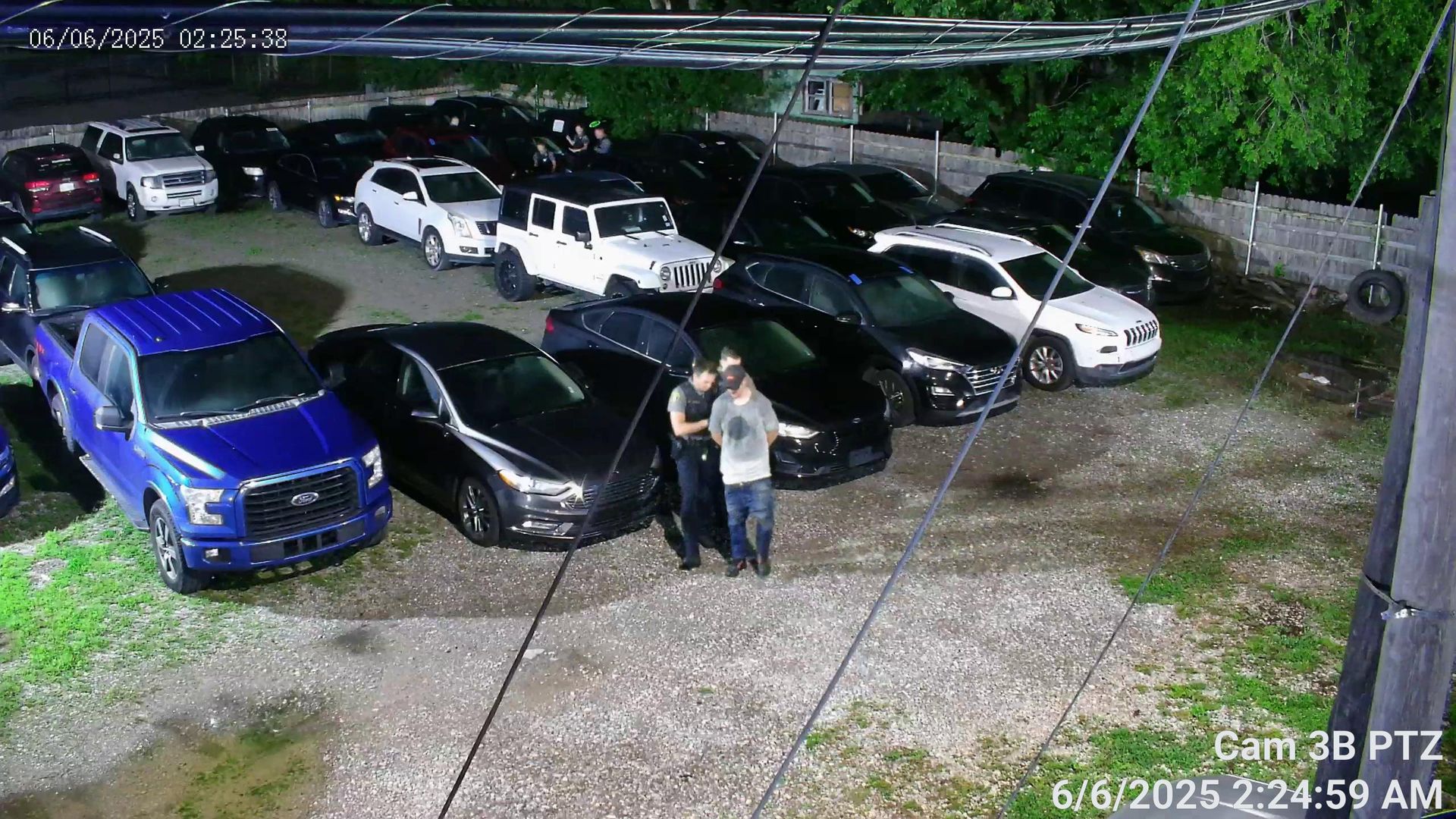 Nighttime parking lot with several parked cars and two people standing near the center under security camera view.