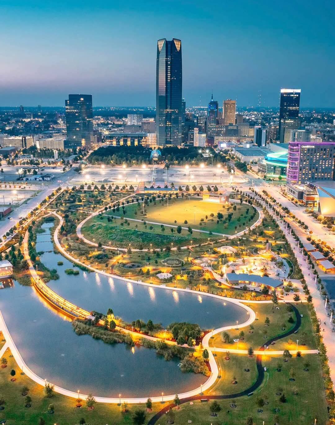 Aerial view of a lit-up circular park and river at dusk with a city skyline in the background