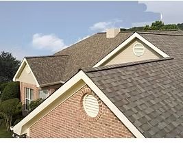 Brown shingled roof with a chimney and gabled dormers on a brick house against a blue sky.