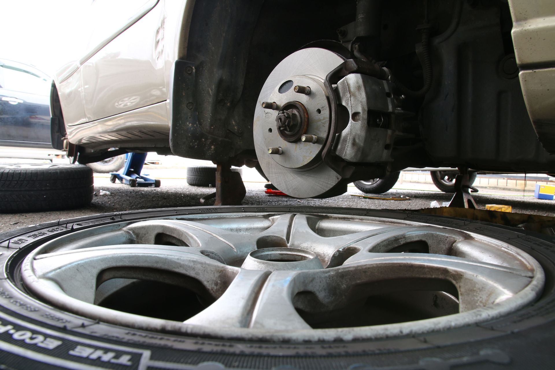 Car wheel removed, revealing brake system, with another wheel in foreground.
