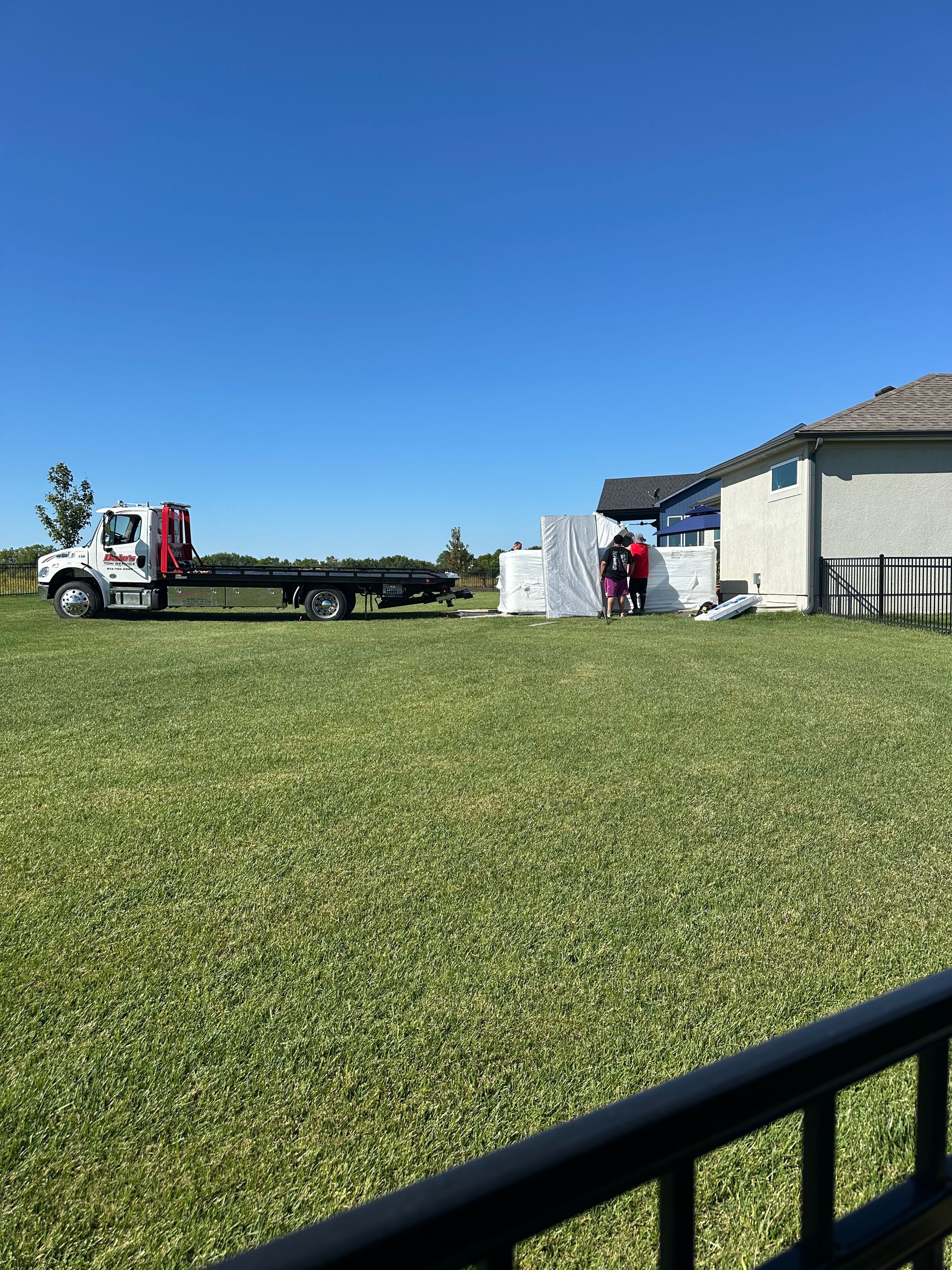 Truck delivering a large, wrapped item to a house with workers present, on a grassy lawn under a blue sky.