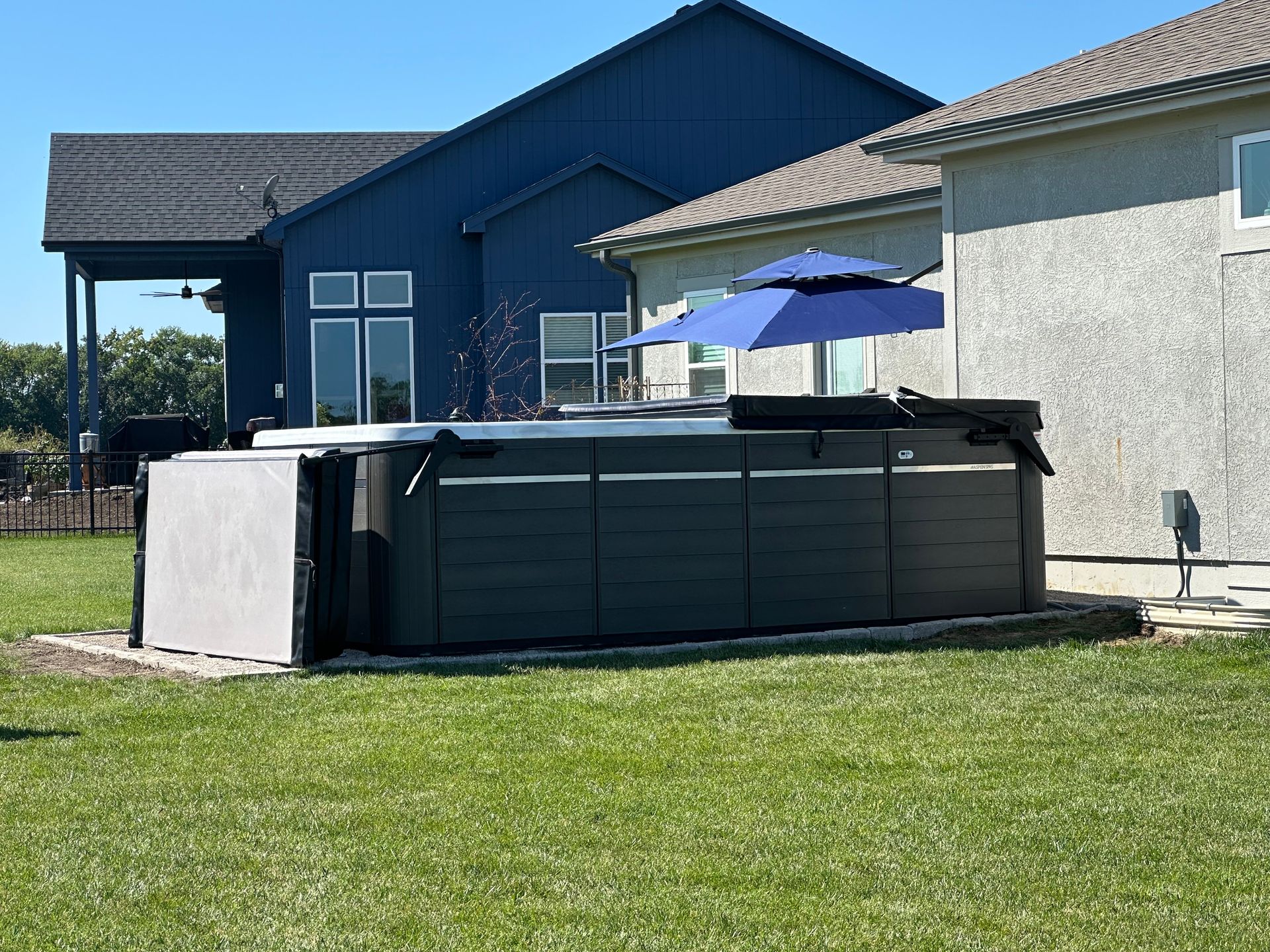 A dark gray hot tub with a blue umbrella in a backyard, surrounded by grass and houses.