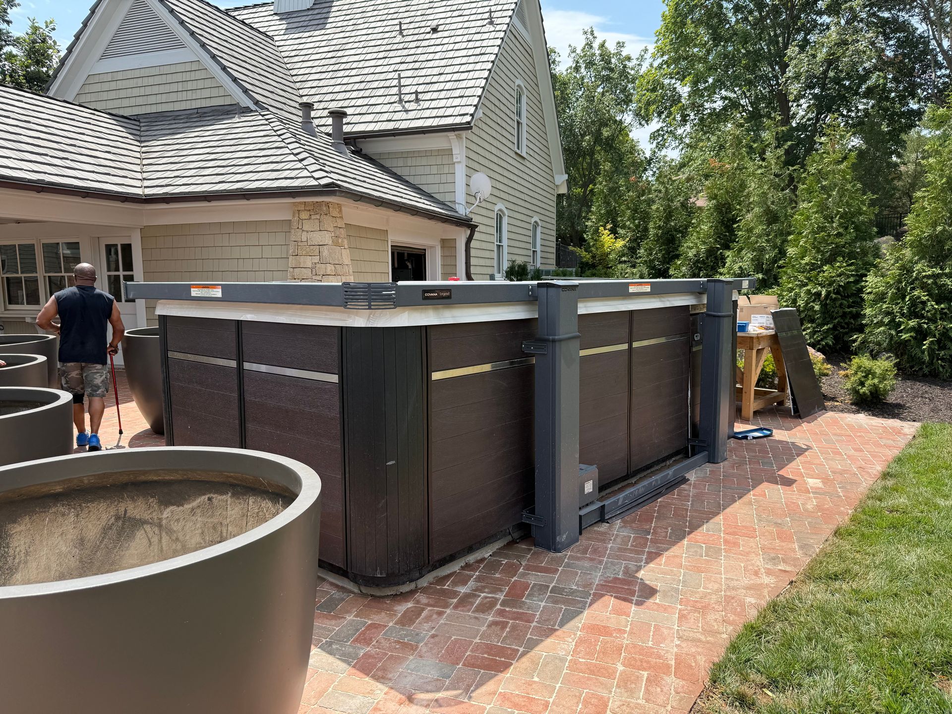 A hot tub with a cover mechanism, set on a brick patio next to a house. A person stands nearby.