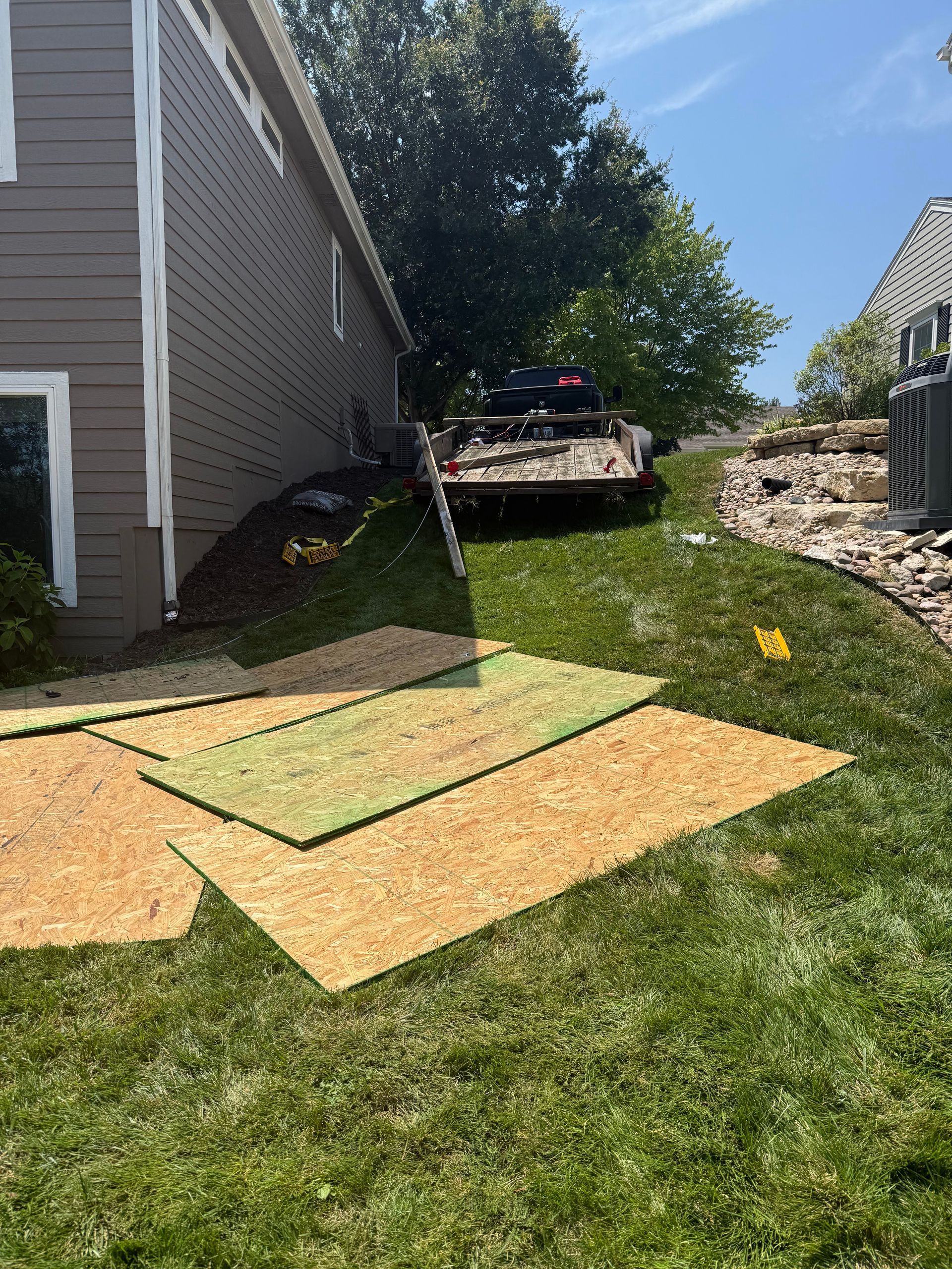 Green grass with wood panels in the foreground. A trailer is parked near a house with a green tree in the background.