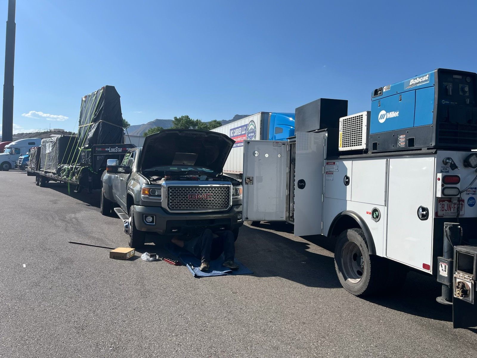 A person works on a black pickup truck with an open hood, parked next to a white service truck on a sunny day.