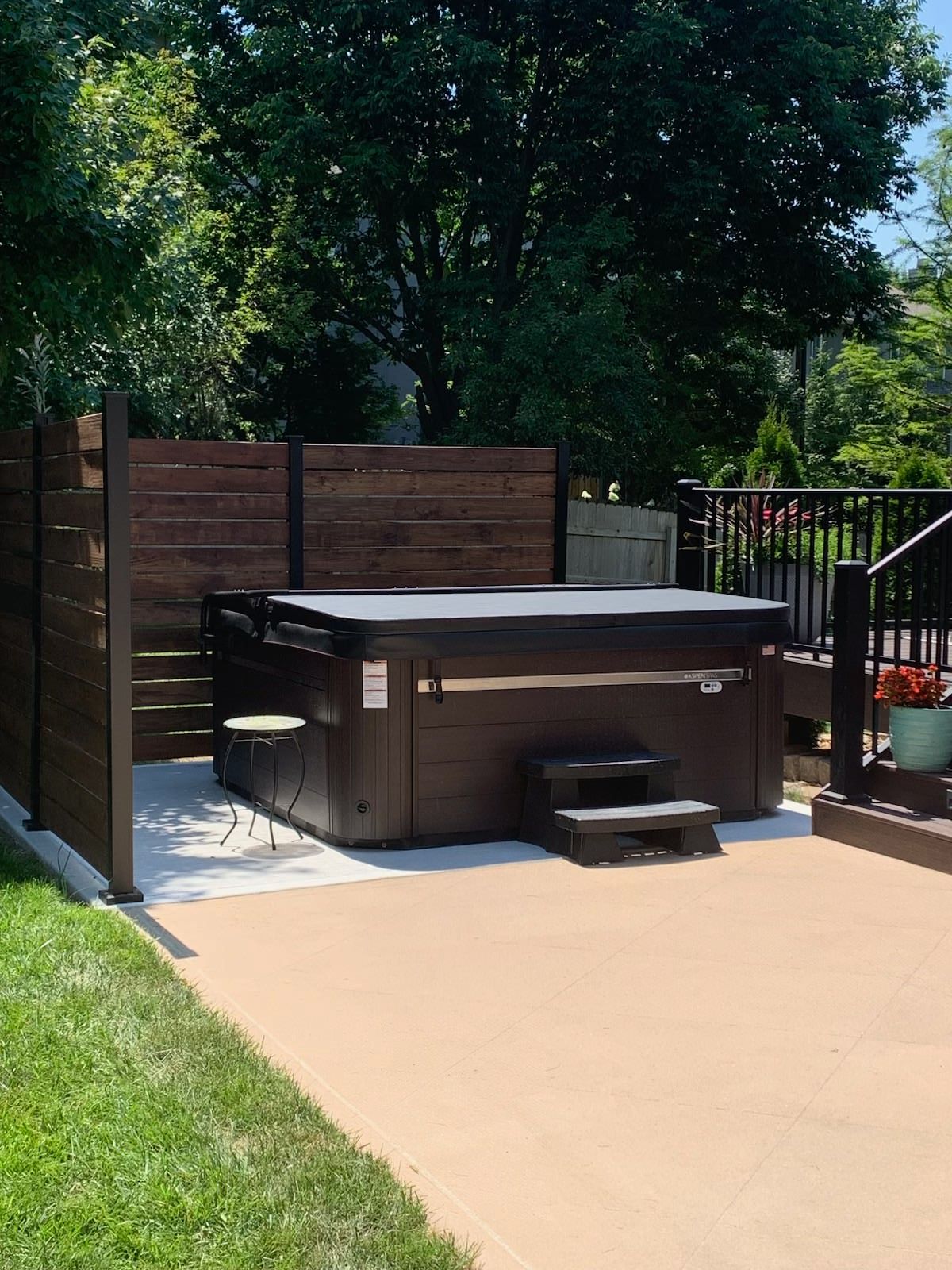 Hot tub on a concrete patio, next to a wood privacy fence and small deck with black railing.