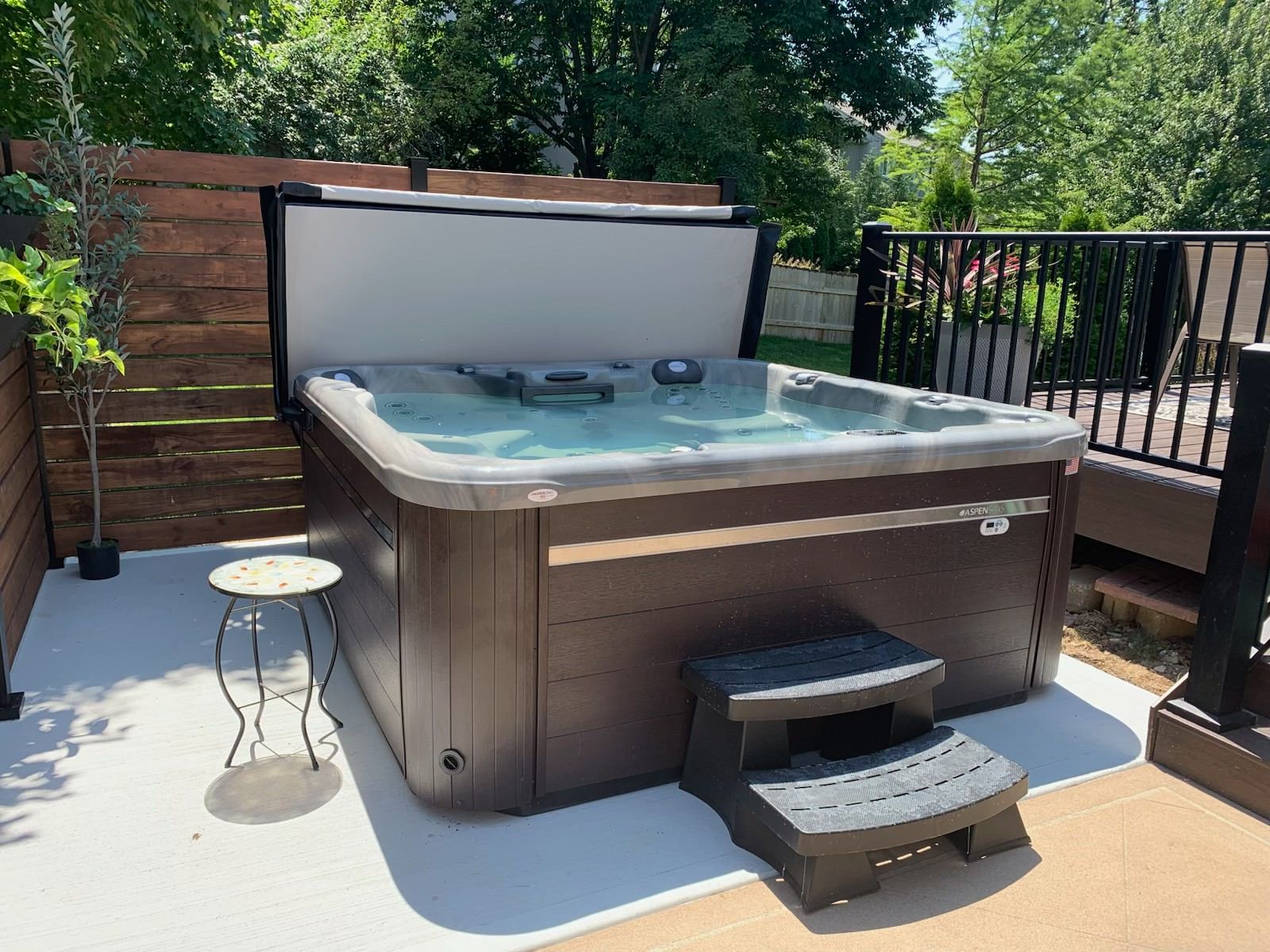 Hot tub with open cover on a concrete patio, next to wooden fence and small step.