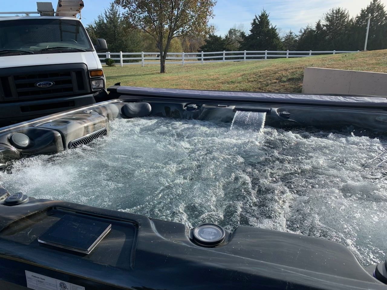Bubbling hot tub with a small waterfall, outdoors near a white fence and a van.