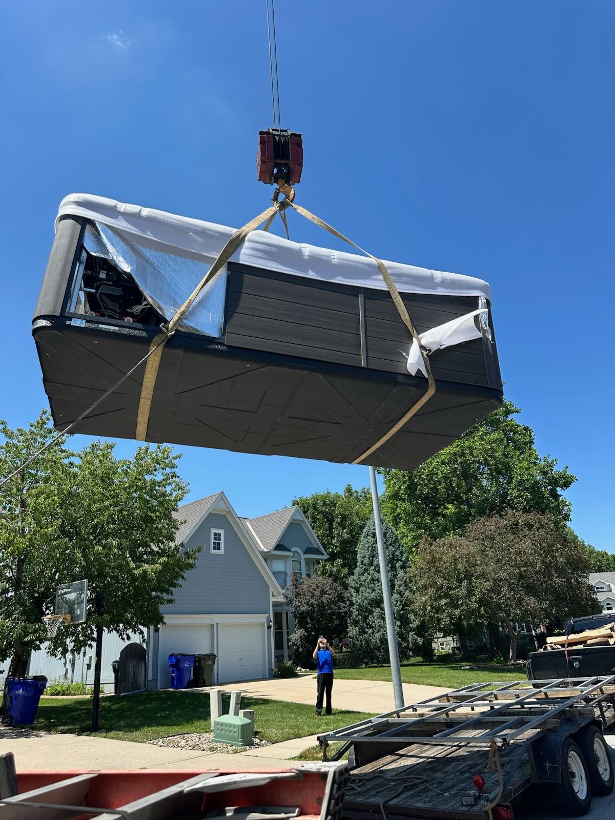 A crane lifting a dark hot tub above a trailer in front of a house, sunny day.