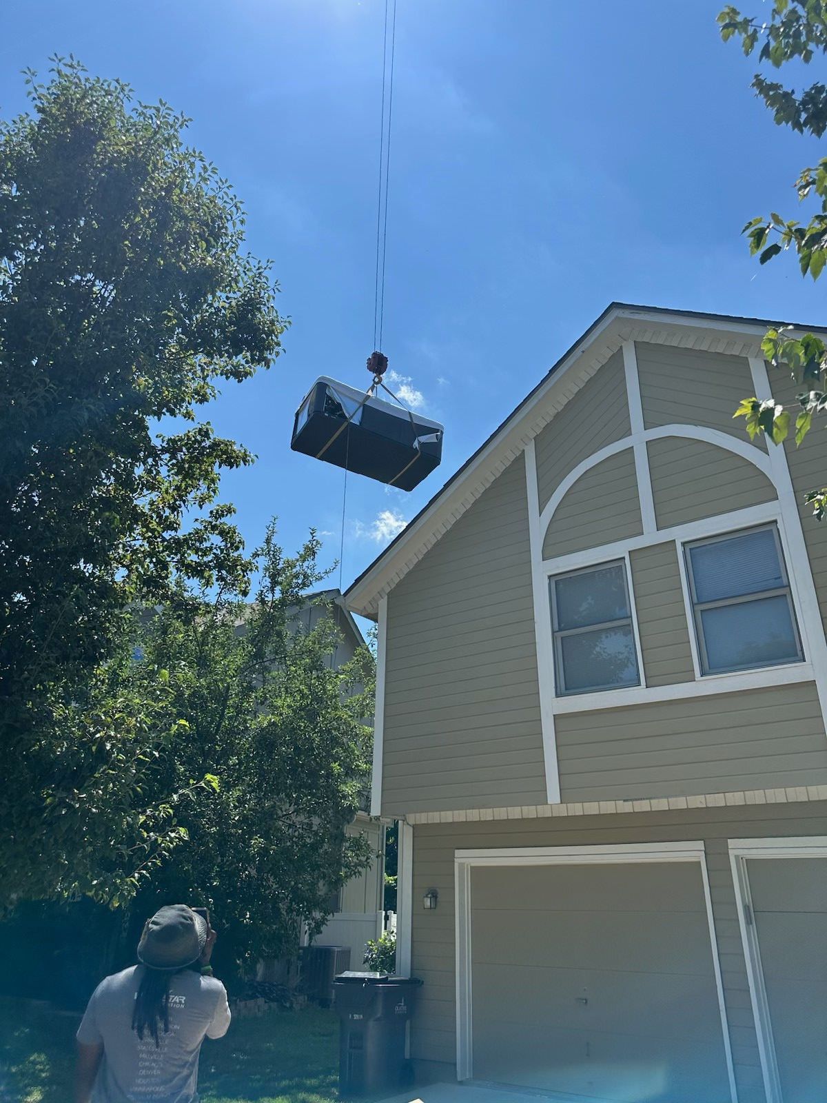 A large box suspended by a cable being lifted over a house on a sunny day. A person looks up.