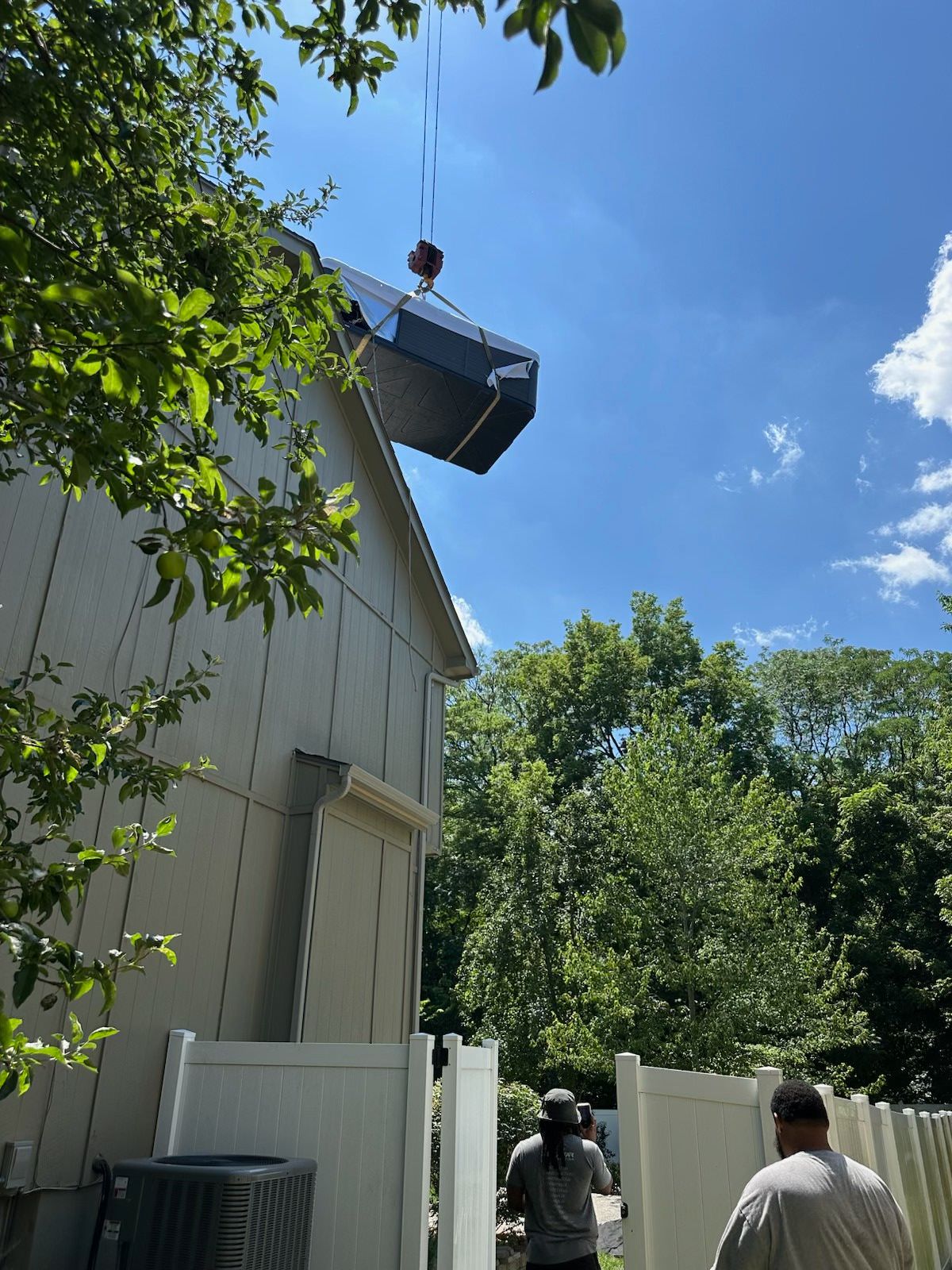 Crane lifting an HVAC unit onto the roof of a house, two workers below. Bright sunny day.