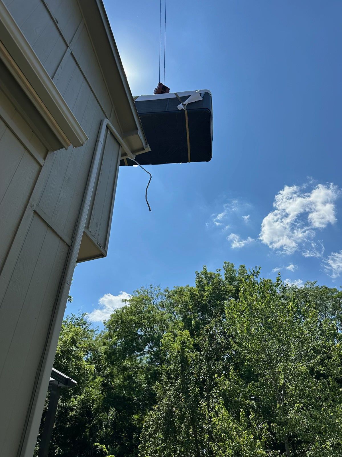 Large dark object being lifted by a crane near a house's roof. Clear blue sky, green trees in the background.