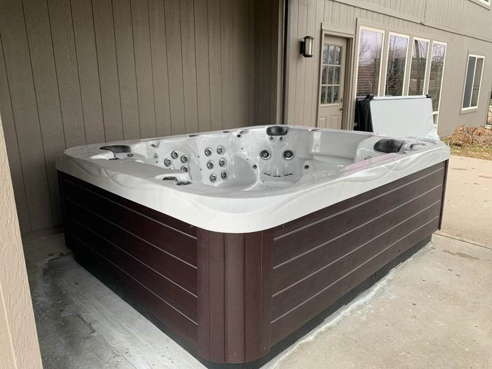 Hot tub, dark brown siding, on a patio next to a house with windows.
