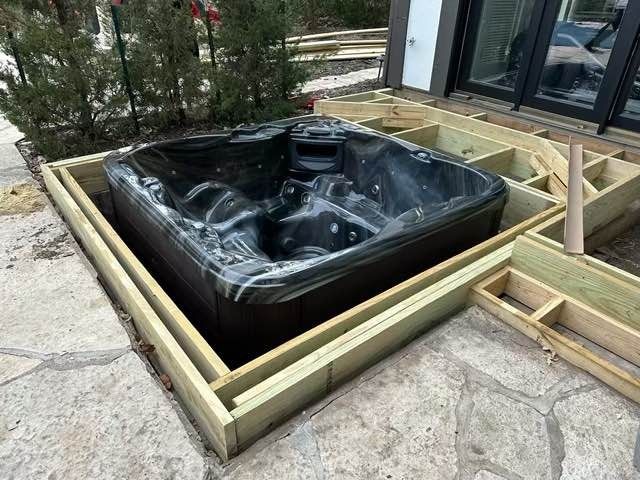 Hot tub installed within a wooden deck frame, on a stone patio, near a building.