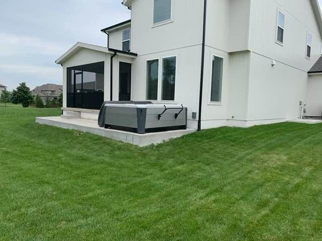 A hot tub sits on a concrete pad next to a white house with a screened porch on a grassy lawn.